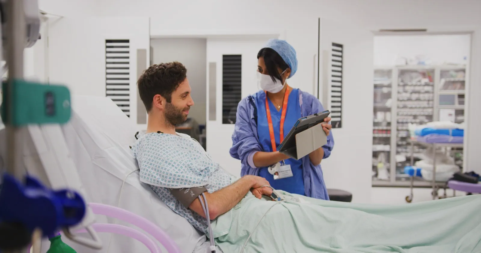 A theatre nurse holding a tablet at the bedside, talking with a patient in a gown before his procedure.