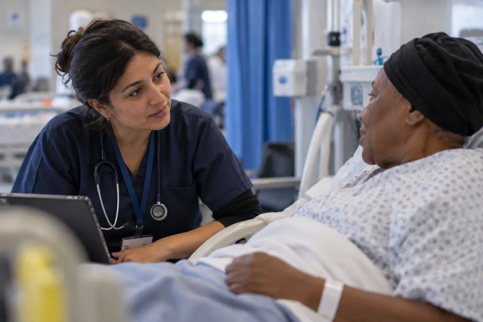 A nurse in scrubs leaning in at a hospital bedside, talking with an older woman sitting up in bed, a tablet resting on the covers between them.