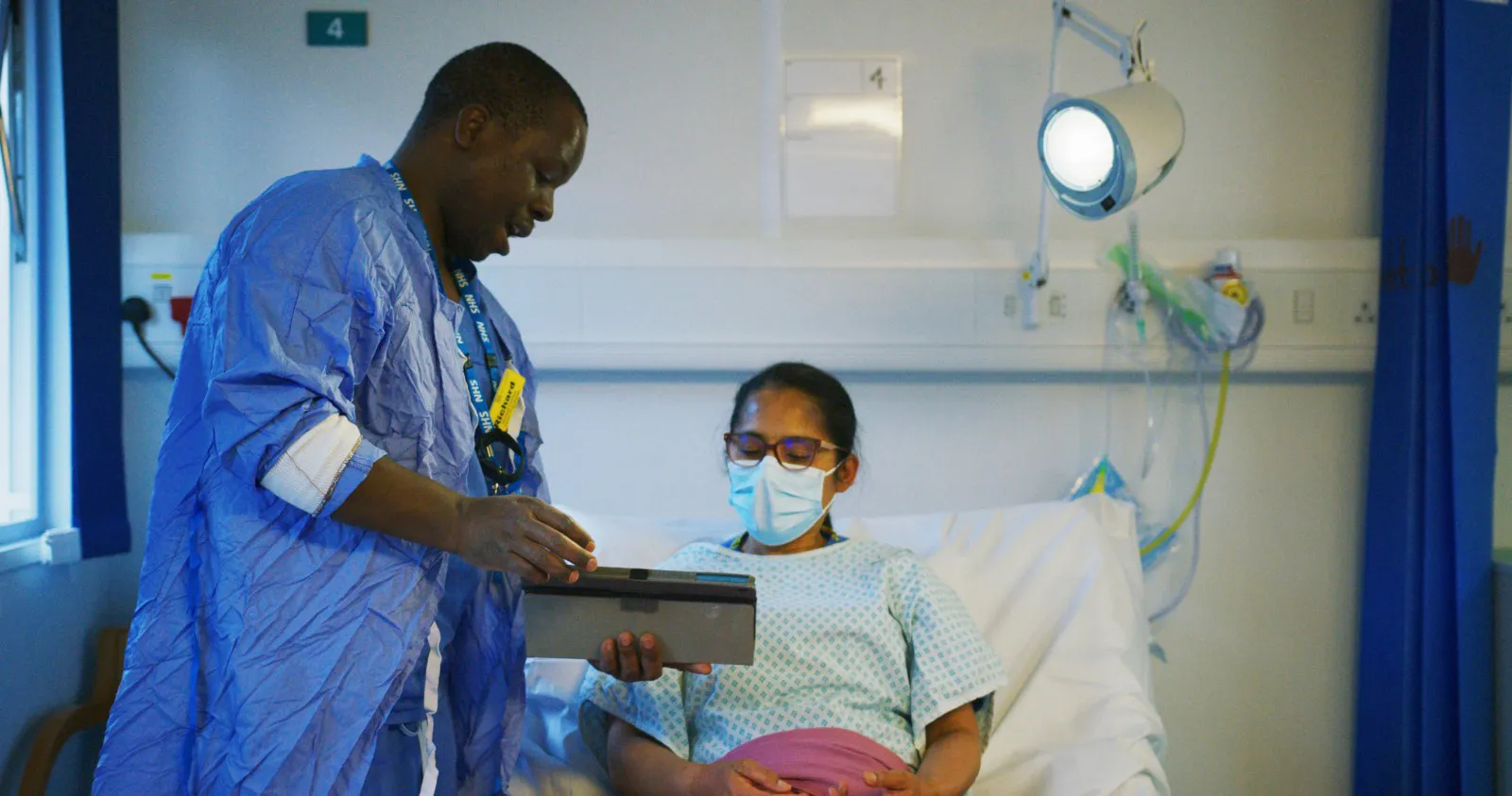 A clinician in NHS scrubs showing CardMedic on a tablet to a pregnant patient sitting up in a hospital bed.
